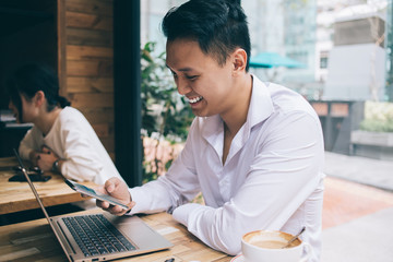 Asian smiling young man sitting at desk with laptop and using smartphone