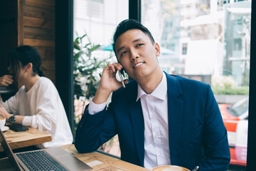 Young Asian smiling businessman sitting and talking on phone against windows