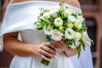 Obraz na płótnie Canvas White wedding bouquet of different flowers and dried flowers in the hands of the bride on a green background. Hands of the bride and dress are artistically blurred against the backdrop