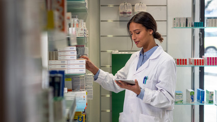An young female pharmacist consultant is checking with a tablet medicines on the shelf of drug...