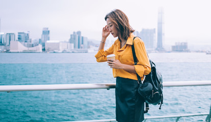 Thoughtful female tourist with coffee to go in hand pondering on vacations near river in Hong Kong, attractive hipster woman trendy dressed thinking on discover metropolis during Chinese holidays