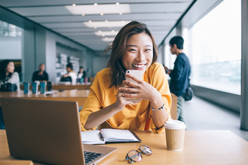 Portrait of cheerful Asian female blogger with cellphone gadget in hands smiling at camera during e learning time, successful Chinese woman rejoicing near desktop with modern laptop computer