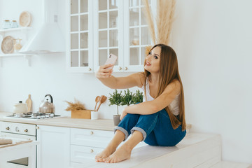 portrait Beautiful young woman in jeans and a T-shirt with a mobile phone takes a selfie in a modern bright Scandinavian kitchen