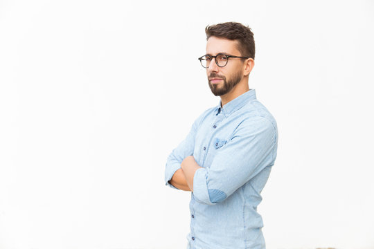 Serious suspicious guy with arms crossed looking at camera. Handsome young man in casual shirt and glasses standing isolated over white background. Male portrait concept
