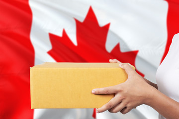 Canada delivery service. International shipment theme. Woman courier hand holding brown box isolated on national flag background.