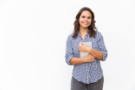Joyful positive student girl holding tablet and smiling at camera. Young woman in casual checked shirt standing isolated over white background. Communication or advertising concept