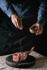 Man's hands sprinkled with salt raw uncooked black angus beef tomahawk steak on bones on linen...