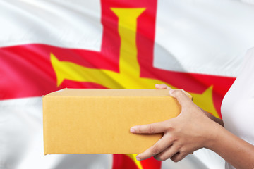 Guernsey delivery service. International shipment theme. Woman courier hand holding brown box isolated on national flag background.