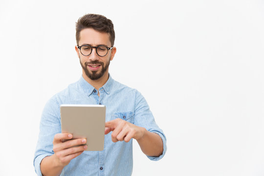 Focused Positive Guy Watching Content On Tablet. Handsome Young Man In Casual Shirt And Glasses Standing Isolated Over White Background. Digital Technology Concept