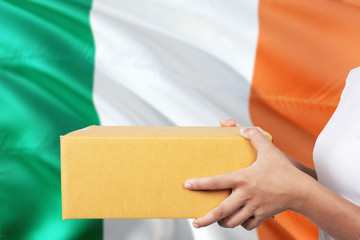 Ireland delivery service. International shipment theme. Woman courier hand holding brown box isolated on national flag background.