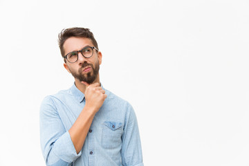Thoughtful male customer thinking hard, looking away at copy space, leaning chin on hand. Handsome young man in casual shirt and glasses standing isolated over white background. Special offer concept