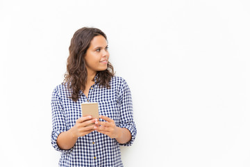 Positive pensive Latin woman with cellphone looking at copy space near her. Young woman in casual checked shirt standing isolated over white background. Communication concept