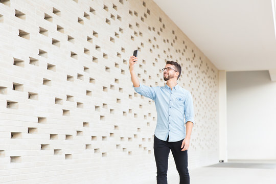 Happy joyful guy with smartphone taking picture or selfie in contemporary interior. Handsome young man in casual denim shirt standing indoors at modern brick wall. Taking picture concept