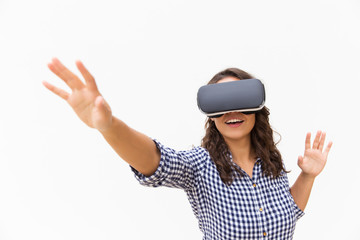 Positive female user in VR goggles touching air and smiling. Young woman in virtual reality headset standing isolated over white background. Augmented reality concept