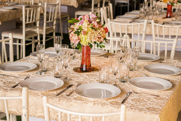 Elegant white table for dinner, with glass cups and vases with roses. With Plates