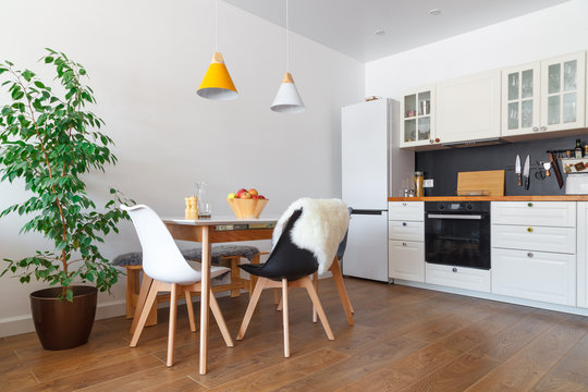 Modern interior of cozy kitchen, dining room, white furniture, wall, black accessories, natural materials, wooden chairs, green flower in pot. Concept scandinavian design apartment of young family
