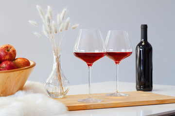 Dark bottle and two glasses of red wine, wooden bowl with apples, decor vase on table in modern...