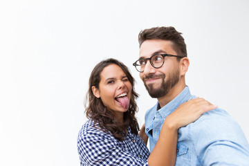 Joyful funny couple grimacing for selfie, smiling and sticking tongue out. Young woman in casual and man in glasses standing isolated over white background. Couple having fun concept