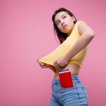 foreign passport with a ticket sticks out of the pocket of the jeans of a young girl who is going on a trip in a yellow T-shirt on a pink background