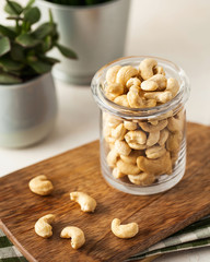 A lot of cashew nuts in a glass container on a wooden Board on a light background. In the background, green plants in pots.