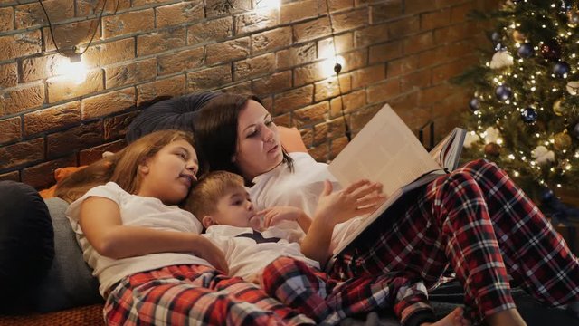 Mom reads a book to children lying on the bed in the bedroom on Christmas evening. The family is dressed in pajamas, a garland and a Christmas tree in the room