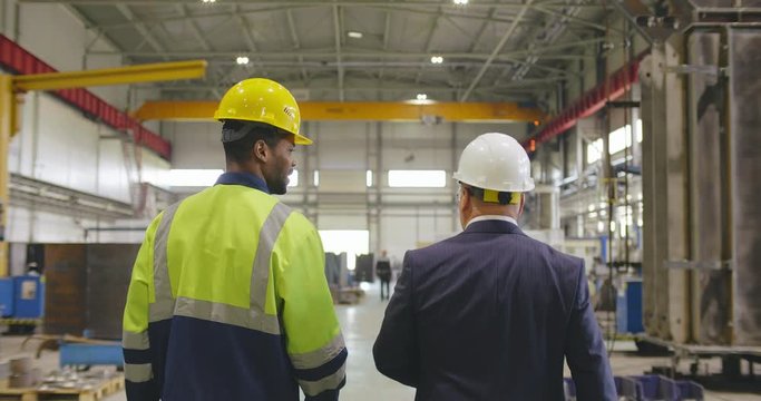 Engineer or businessman and factory worker foreman walking and discussing with tablet pc. Two colleagues in helmets go through industrial background of modern factory.