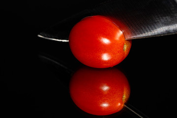 A single grape tomato on a highly reflective surface being sliced by a knife on a black background with vibrant colors and high detail. Room for copy.