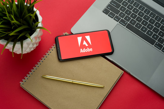 Tula, Russia, november 26, 2019: Adobe logo on the smartphone screen is placed on the Apple macbook keyboard on red desk background.
