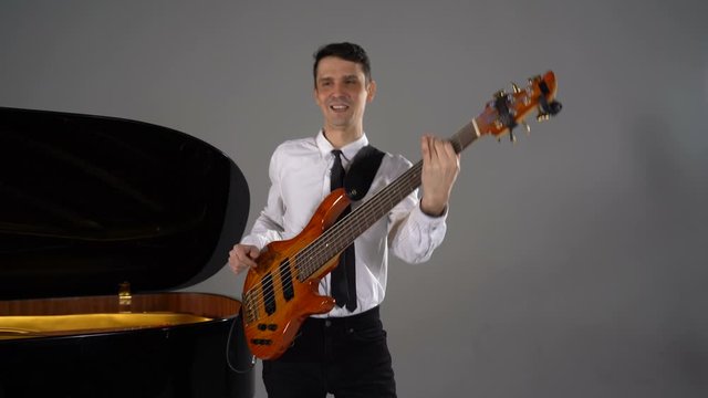Male musician plays bass guitar on grey background next to piano