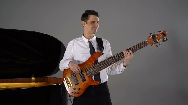 Male musician plays bass guitar on grey background next to piano