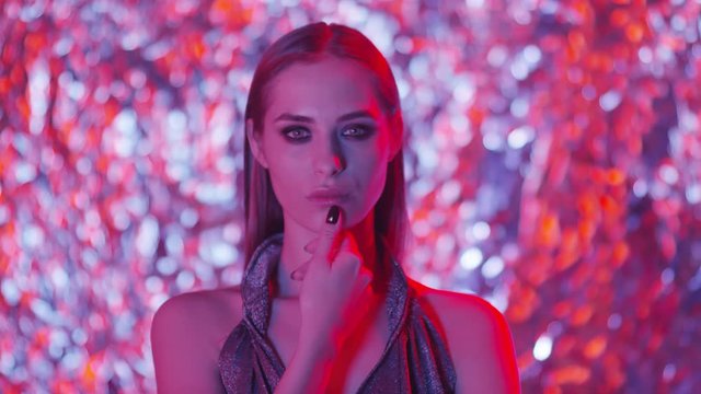 Chest up shot of young beautiful model with smoky eyes makeup holding hand on chin and looking at camera while posing against shining background in studio with neon light