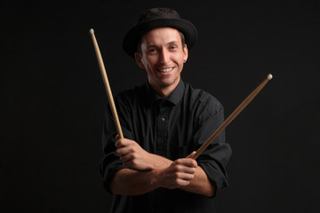 Stylish man drummer in a black shirt and hat playing drums with sticks over dark background.