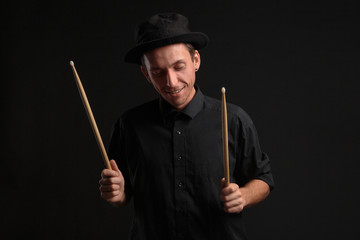 Stylish man drummer in a black shirt and hat playing drums with sticks over dark background.