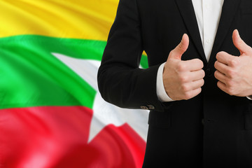 Myanmar businessman showing thumbs up behind country flag with copy space. Successful international...