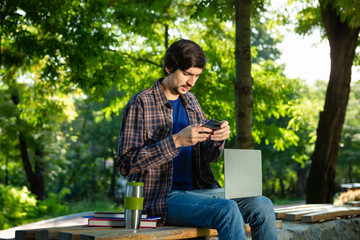 Young brunet freelancer with beard and mustache sitting with a laptop and coffee in a park.