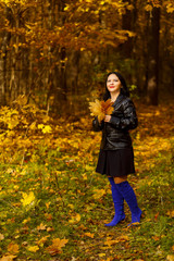 A young brunette woman in blue boots is walking in an autumn park with yellow leaves in her hands