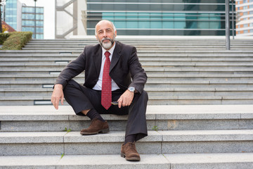 Positive mature company owner holding cellphone and enjoying work break outside. Elderly man in formal suit and tie sitting on outdoor office stairs. Business leader outside concept