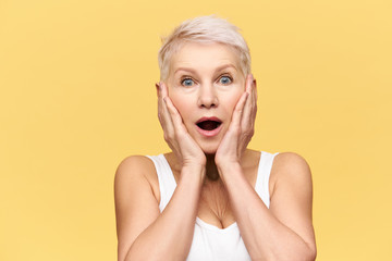 Studio shot of emotional excited middle aged Caucasian woman with blonde pixie hairstyle posing isolated in white tank top holding hands on her face, expressing amazement and full disbelief
