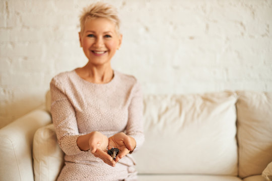 People, business, real estate and immovable property concept. Picture of happy attractive blonde mature female realtor holding keys to your new apartment, smiling, sitting on couch. Selective focus