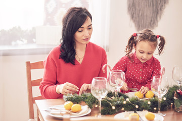 Cheerful mother and her daughter laughing, having fun with Christmas tree and star shaped cookies at the kitchen table. They are in red clothes wearing reindeer antlers and Santa hat headbands.