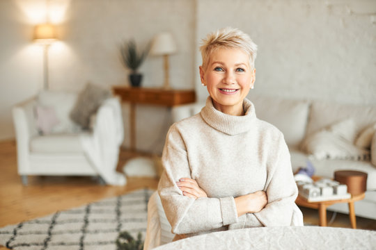 People, Coziness, Domesticity And Season Concept. Charming Beautiful Retired Woman Spending Leisure Time Indoors At Home Looking At Camera With Confident Smile, Keeping Arms Folded On Her Chest