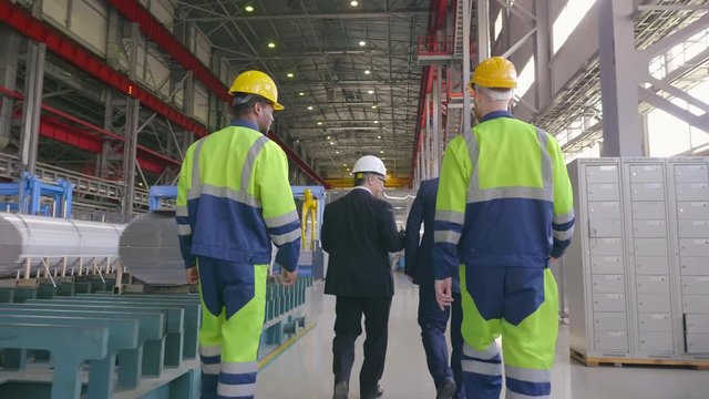 Two businessmen visiting inspecting warehouse followed by workers. Team of engineers in hard hat walking through facility of modern heavy industry factory