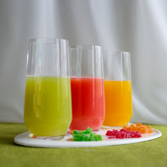 Three clear glasses of multicolored juice, red, green and yellow, with slices of dried mango, kiwi and orange next to them. Healthy diet