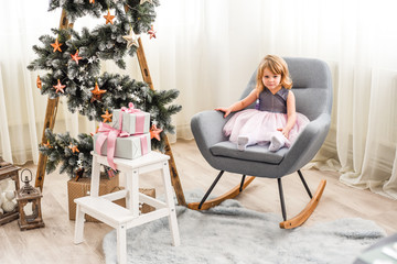Fototapeta premium beautiful girl in a pink dress sitting on a chair near a Christmas tree