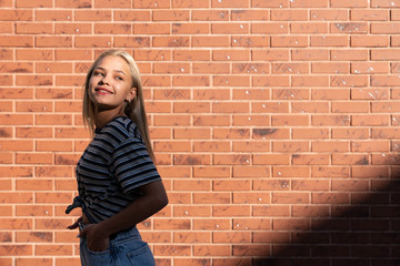 Attractive young blond girl in grey striped t-shirt smiling and looking over her shoulder at right side, isolated over red brick wall
