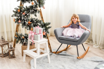 Fototapeta premium beautiful girl in a pink dress sitting on a chair near a Christmas tree