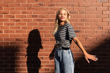 Attractive blonde teen girl in grey t-shirt and jeans dancing in the sun isolated over red brick wall