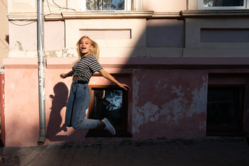 Happy excited blonde teen girl with loose hair dressed in casual clothes jumping among the street of the old city
