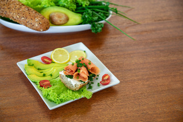 A sandwich with salmon, cream cheese, homemade bread and parsley on a white plate with avocado, salad, lemon and cherry tomato, wooden table