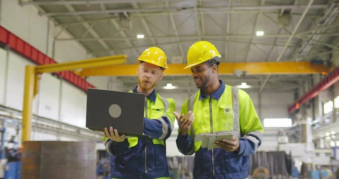 Two factory workers walking with discussion holding tablet pc. Industrial heavy industry enterprise background. Foremen in helmets and uniform walking through factory facility